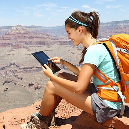 A young woman hiking on top of a mountain sits down as she views her Quorum account on her laptop.