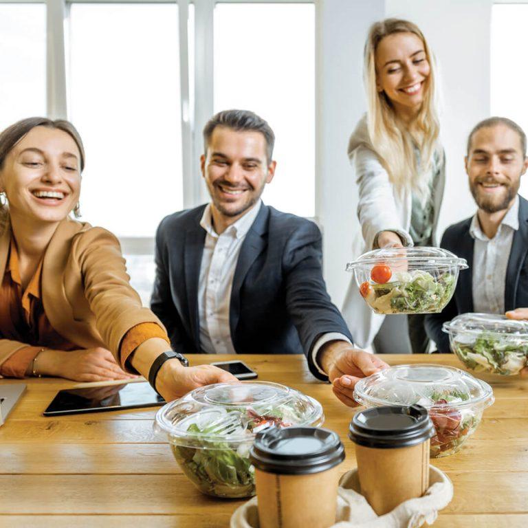 Smiling co-workers grab salad lunch orders from middle of conference room table.