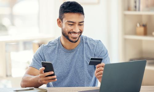 Smiling man holds credit card in one hand, phone in the other, with laptop on desk.