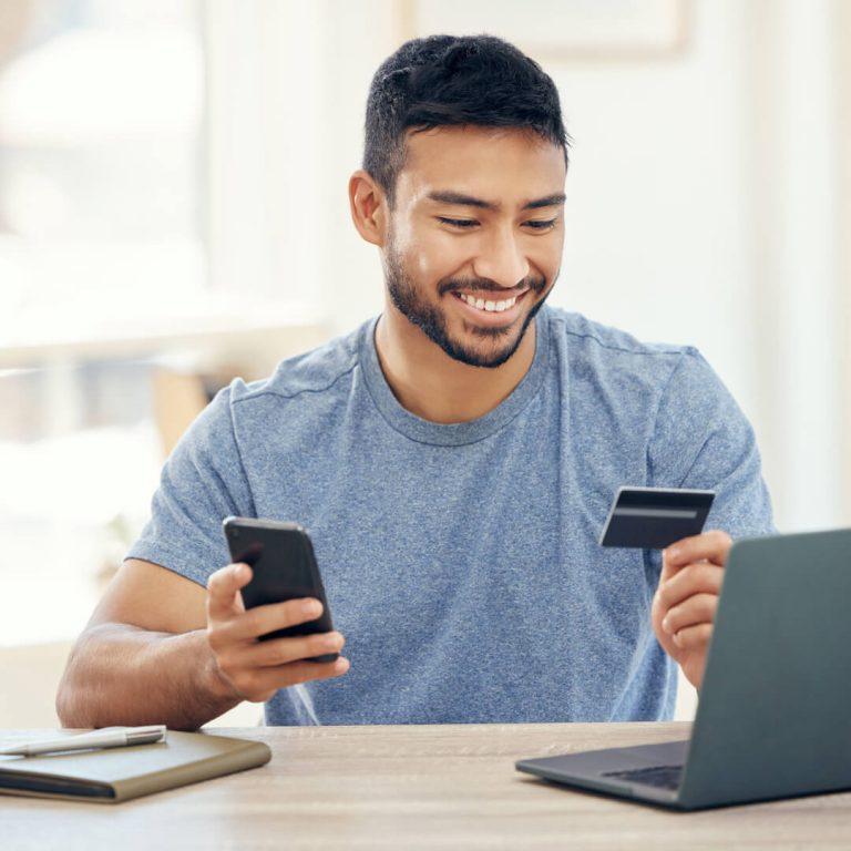 Smiling man holds credit card in one hand, phone in the other, with laptop on desk.