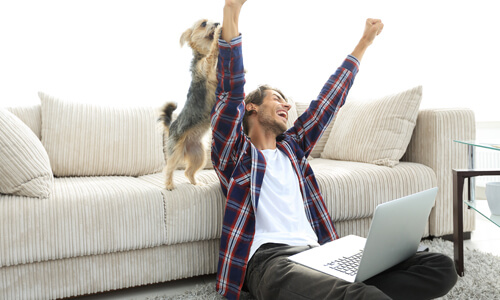 Man on floor with laptop, cheering with hands in the air; his dog behind him, also cheering.