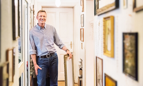 Man smiling in hallway with his private art collection framed and displayed on wall.