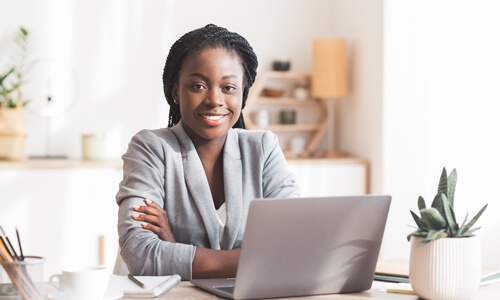 Businesswoman seated behind desk, looking out from behind her laptop, smiling confidently at camera.