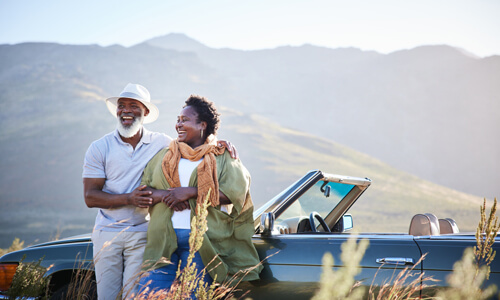 Older couple standing outside classic convertible admiring the scenery, mountains in background.