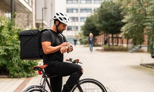Gig worker (delivery man on bicycle) checks food order on his wearable, with food strapped to his back.