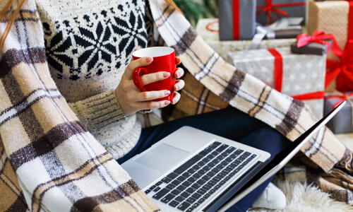 Woman in flannel blanket and holding mug, looking at her laptop.
