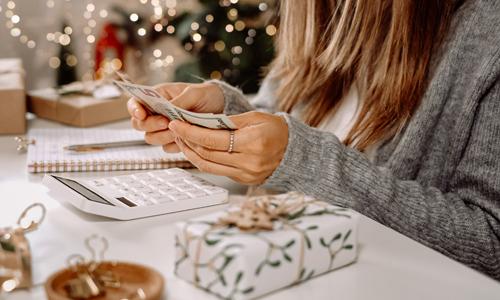 Woman budgeting money for the holidays, holding cash, looking at calculator and near gifts.