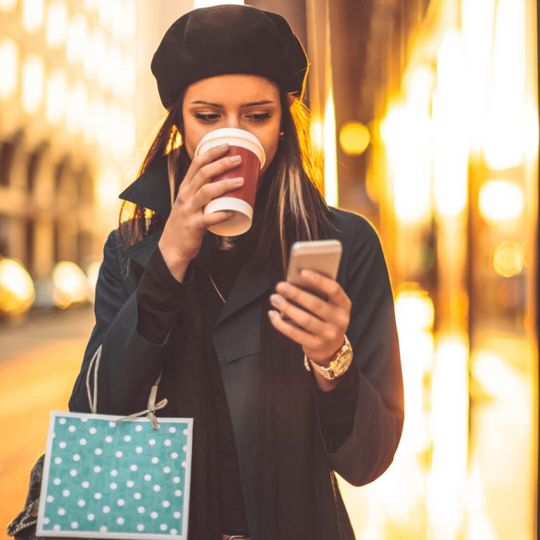 Woman walking down street shopping, drinking coffee, and browsing her phone.