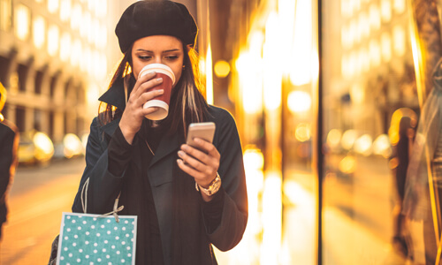 Woman walking down street shopping, drinking coffee, and browsing her phone.