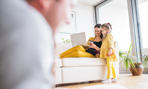 Mother and child in living room; daughter watches her mother purchase something online with a credit card.