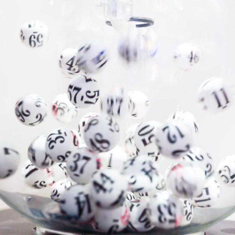 Black and white lottery balls are in motion in a glass bowl that is sitting on a wooden stand. One ball is being sucked up a tube.