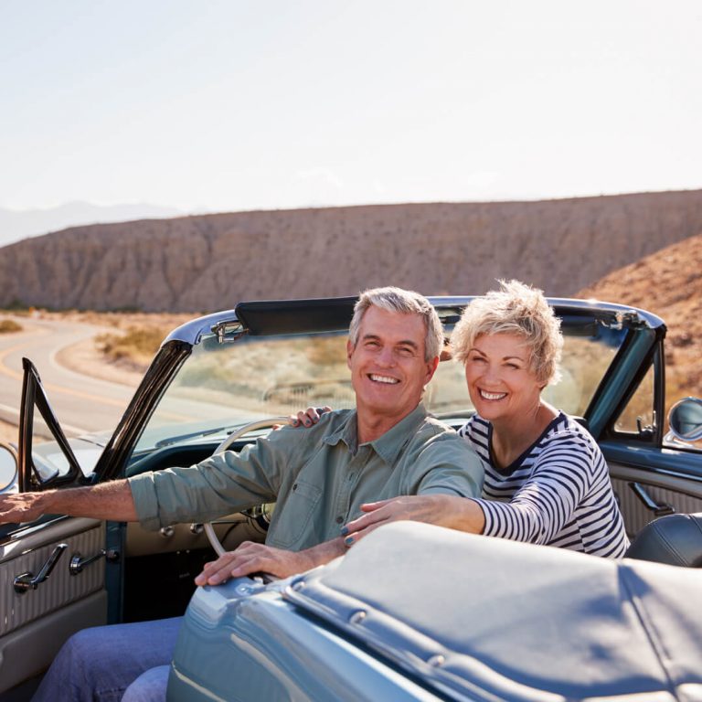 Senior couple smile to camera from parked open top classic car on luxury roadtrip.