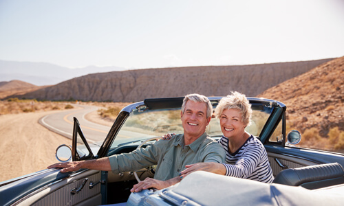 Senior couple smile to camera from parked open top classic car on luxury roadtrip.