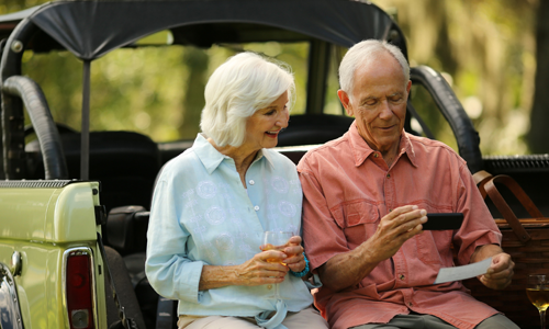 Elderly man and woman stand outside at the back of their truck taking a picture of a check with their mobile device.
