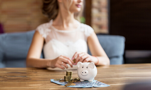 A woman in a bridal gown sitting at a table with money and a piggy bank, wondering how she'll pay for her wedding.