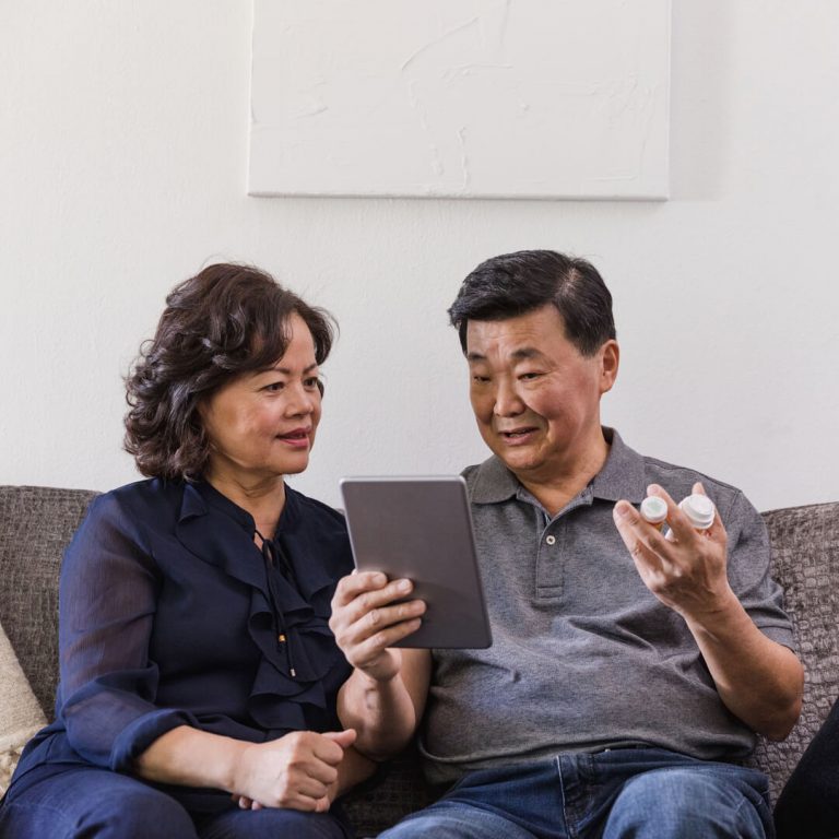 Couple seated on couch with prescription medicine in hand, looking at tablet.