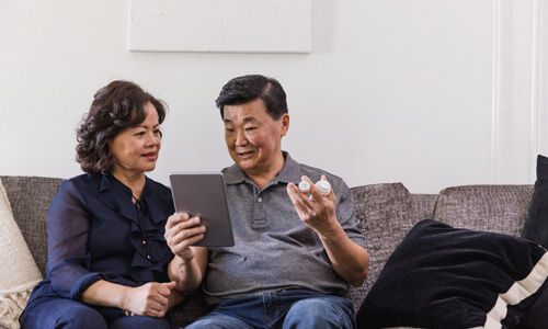 Couple seated on couch with prescription medicine in hand, looking at tablet.