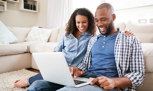 Man and woman seated on couch smiling and looking at their laptop.