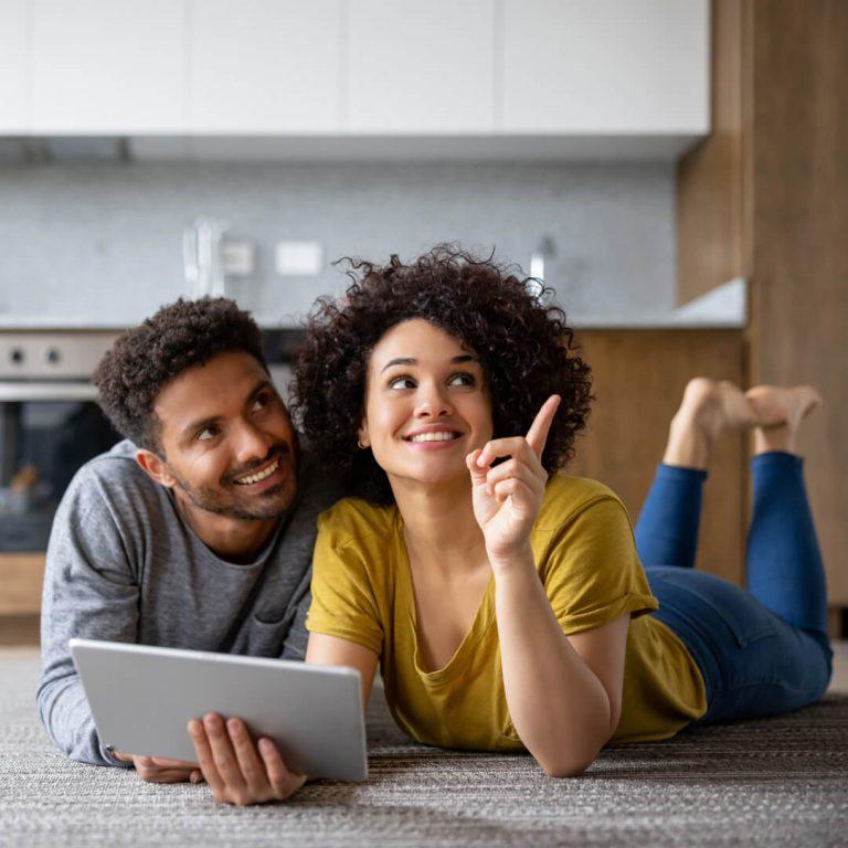 Couple in kitchen, looking at laptop, while pointing out potential renovations, deciding whether to move or improve their home.