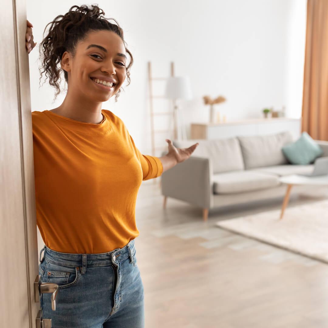 Smiling woman standing at door of new apartment, welcoming you inside.