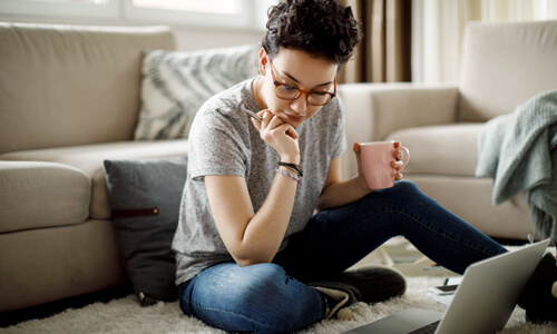 Young woman seated on floor in living room with laptop, reviewing her finances and student loans.