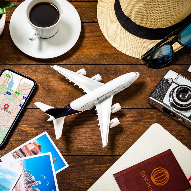 Items on table, representing a summer vacation: passport, phone, camera, sunglasses, hat, model airplane.