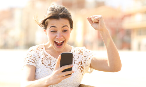 Woman looking at her phone, holding up her hand in excitement after learning her tax refund amount.