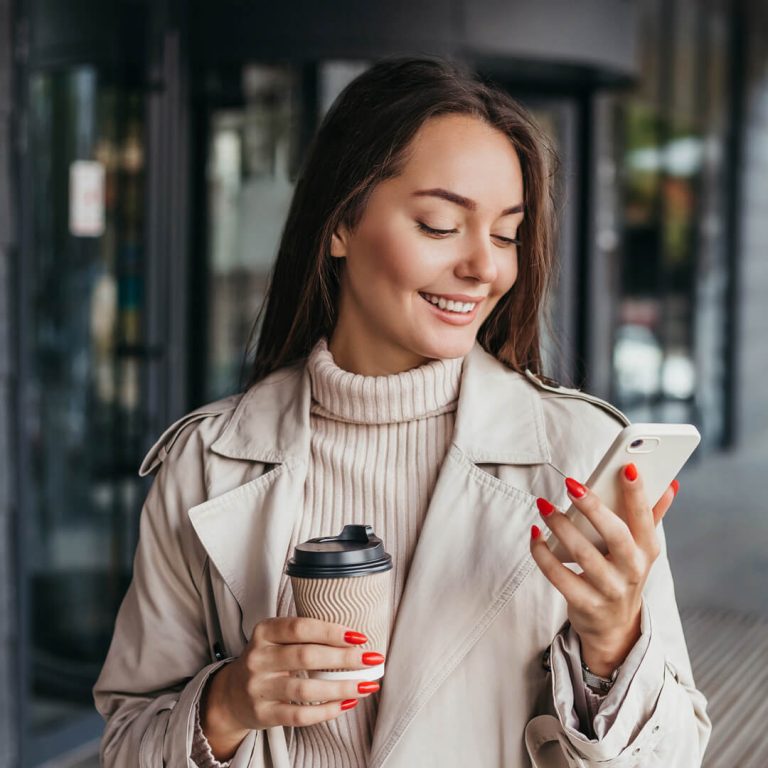 Smiling woman holding coffee and shopping, smiling at her mobile phone, checking her available balance.