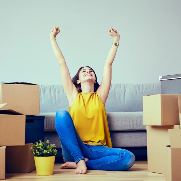Happy young woman sitting on floor surrounded by moving boxes with her hands in air.