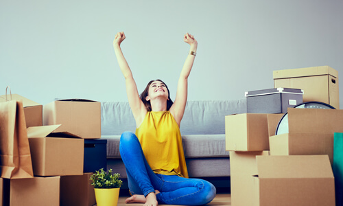 Happy young woman sitting on floor surrounded by moving boxes with her hands in air.