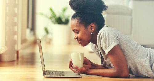 Young woman entrepreneur lying on her stomach on floor with a cup of coffee, looking at her laptop.