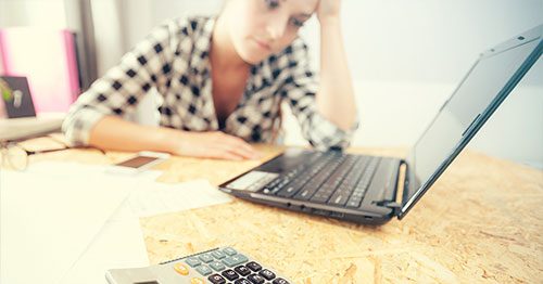 Stressed woman looking at laptop and calculator after incurring unexpected household expenses.