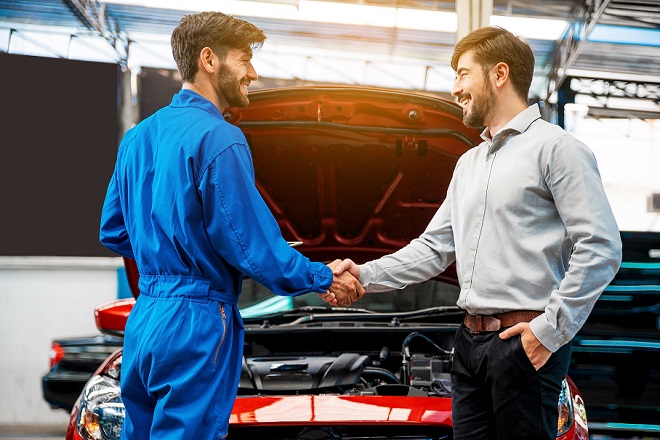mechanic shaking hands with male customer over car with hood open in garage