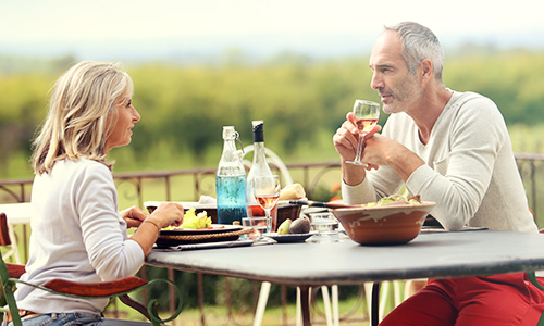 Recent retiree couple sit outside enjoying lunch and wine overlooking a vineyard.