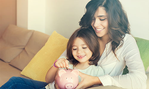 Mother and daughter sitting on couch smiling and adding coins to a piggy bank.