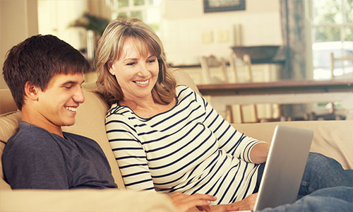 Mom sitting on couch with teenage son, looking at laptop together.