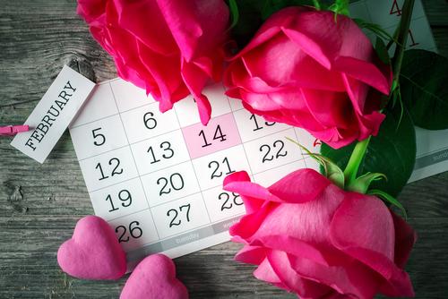 A calendar on a wooden table showing February14th and surrounded by red and pink roses and hearts.