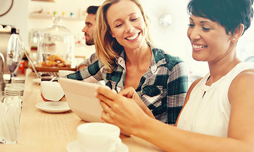 Two 40-year-old women smiling at counter enjoying coffees and looking at laptop.